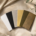 Color swatches in black, white, gold, and brown on a textured beige surface with green leaves.